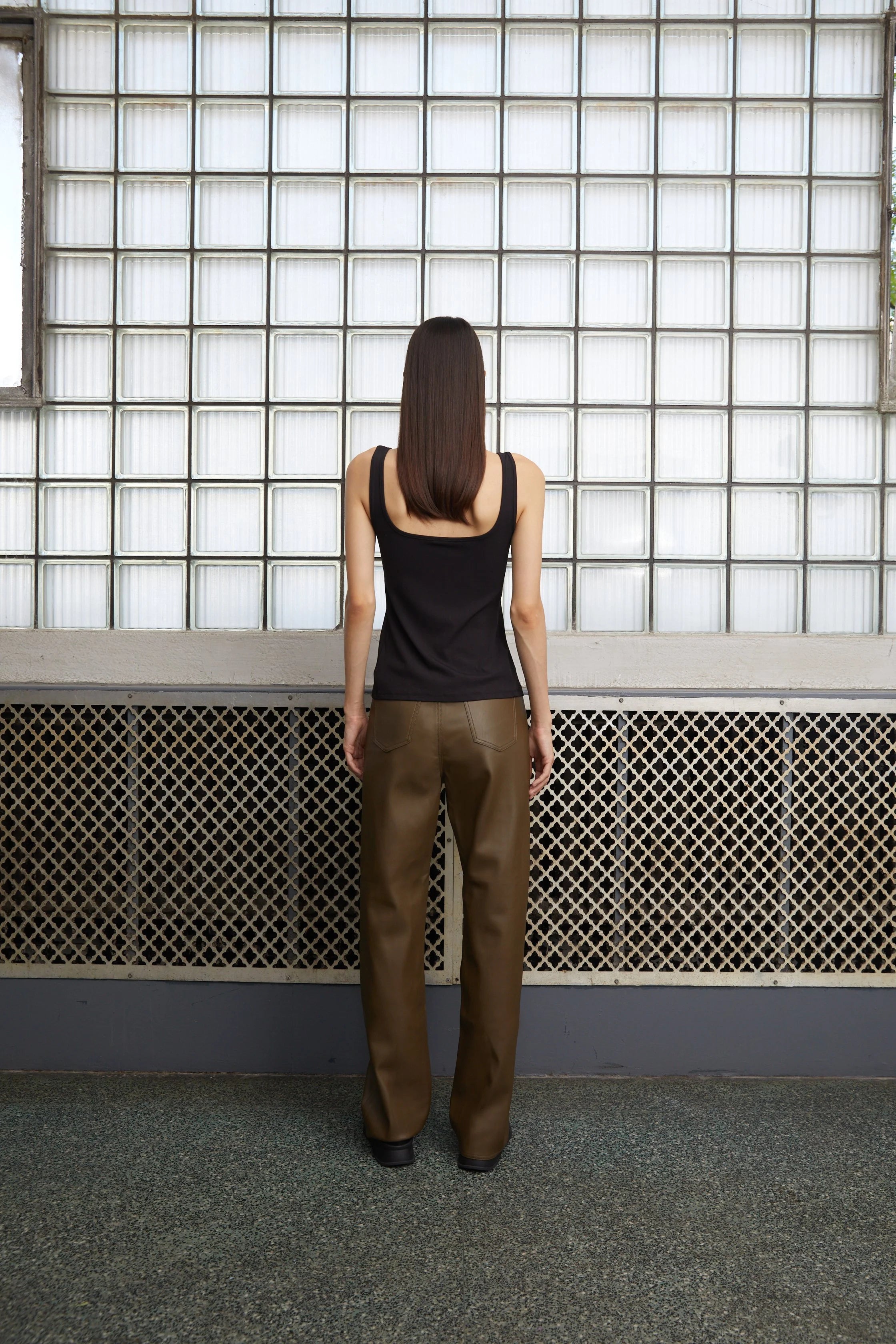 Person wearing a black tank top and brown pants standing in front of a grid-patterned wall.