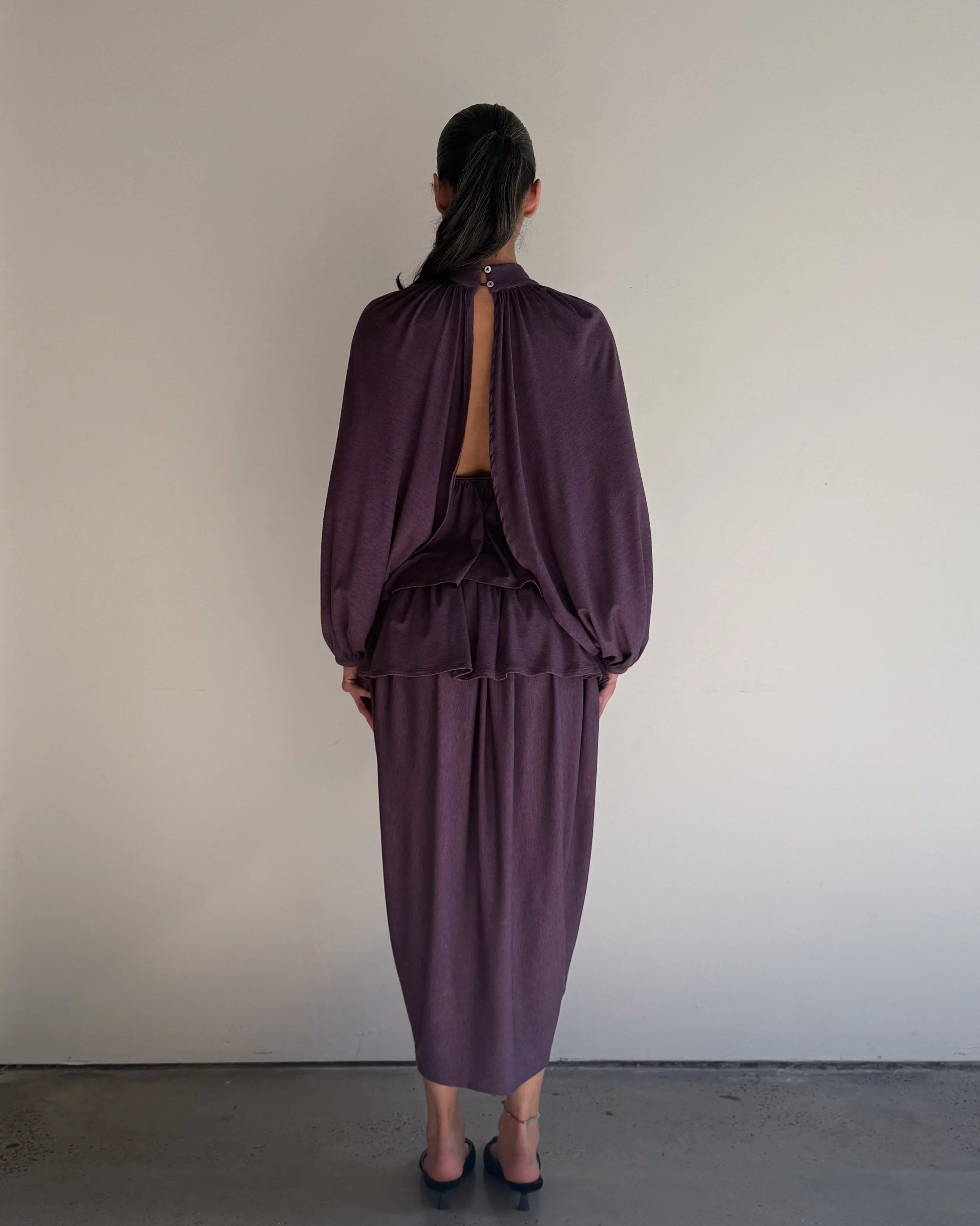 Person wearing a purple outfit standing against a plain wall.
