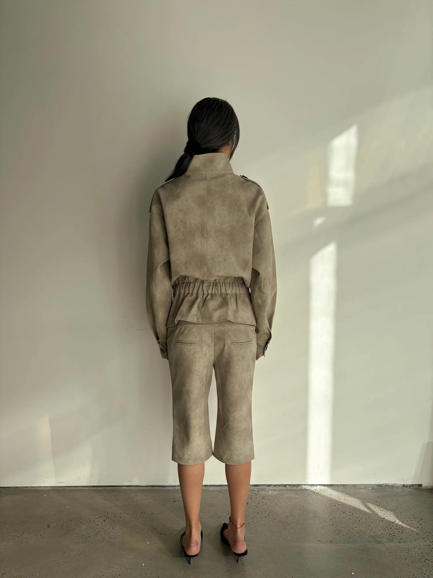 Person wearing a beige outfit standing against a white wall with sunlight casting shadows.