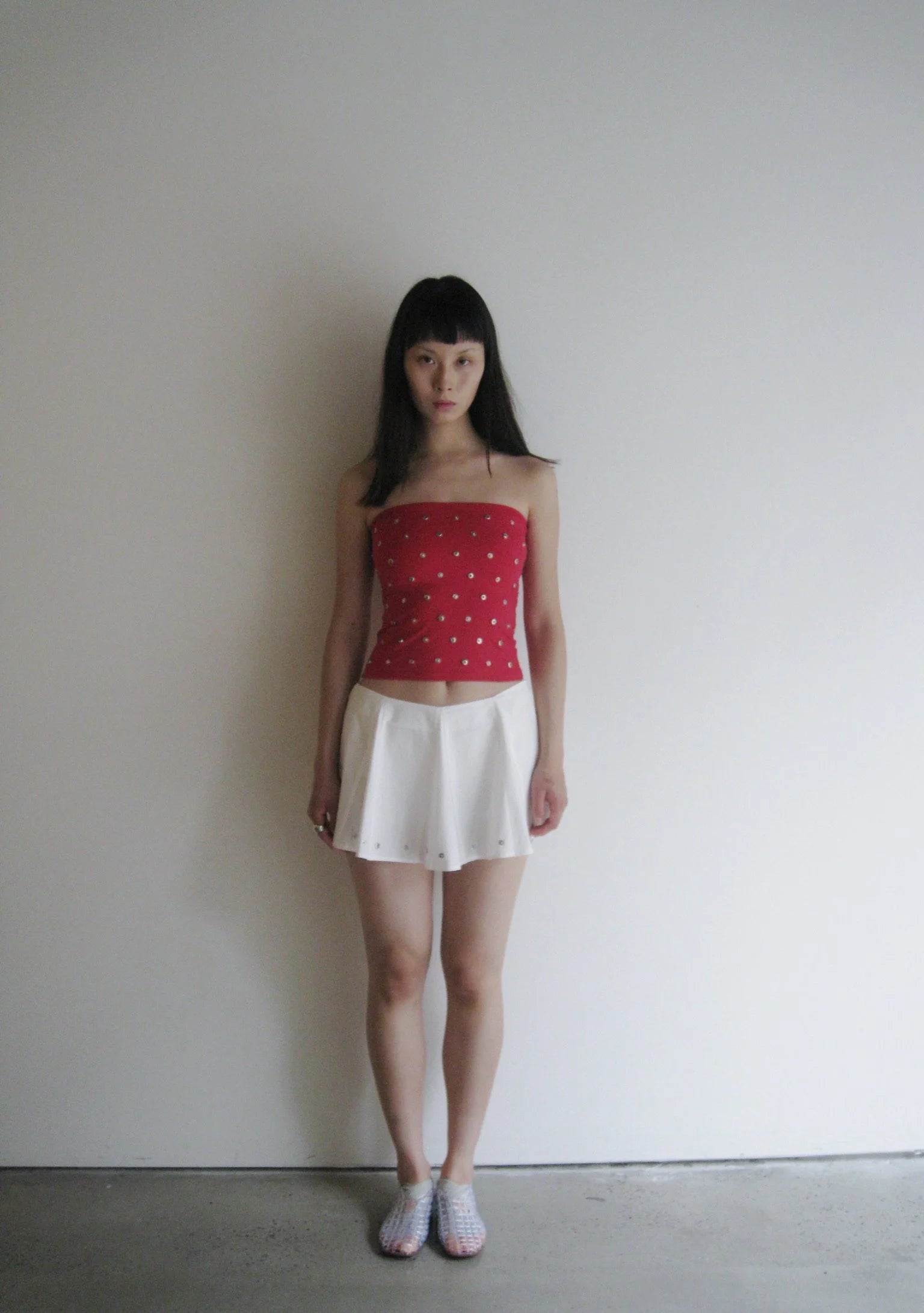 Woman wearing a red strapless top with embellishments and a white skirt against a plain background